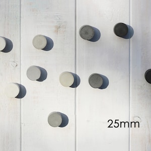 Op de afbeelding: Tien ronde betonnen wandhaken in verschillende grijstinten en zwart, elk ongeveer 2,5 cm in diameter, zijn gerangschikt op een wit houten oppervlak. De tekst "25mm" is zichtbaar.