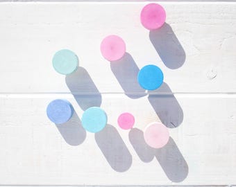Puede incluir: Surtido de pomos redondos de madera de color pastel en tonos rosa, azul y azul claro. Los pomos están dispuestos en un patrón sobre una superficie de madera blanca.