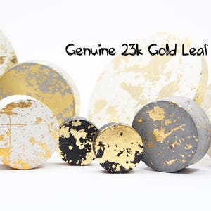 May include: A selection of concrete ornaments in various shapes, including spheres and discs. The objects are painted in white, grey, and black, with genuine 23k gold leaf accents. The text "Genuine 23k Gold Leaf" is visible.