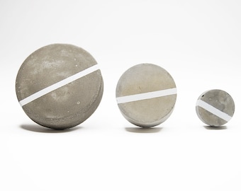 May include: Three round concrete coasters with a white stripe across the top. The coasters are different sizes.