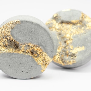 Puede incluir: Dos objetos decorativos redondos grises con detalles de pan de oro. El pan de oro está incrustado en el material gris, creando una apariencia texturizada única. Los objetos están sobre un fondo blanco.