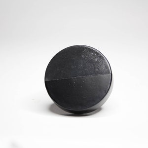 Könnte beinhalten: Ein schwarzes, kreisförmiges Objekt mit strukturierter Oberfläche und einer horizontalen Linie in der Mitte. Das Objekt befindet sich vor einem weißen Hintergrund, wodurch ein kontrastreiches Bild entsteht.