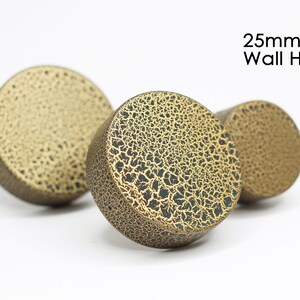 Op de afbeelding: Drie goudkleurige wandhaken met een gekrakelde afwerking. De haken hebben een diameter van 25 mm en zijn ontworpen om items aan een muur te hangen.