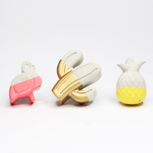 Può includere: Tre piccole figurine di cemento dipinte a tema tropicale. Un fenicottero rosa, un cactus dorato e un ananas giallo.