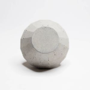 May include: A grey concrete geometric vase with a round opening at the top. The vase has a textured surface.