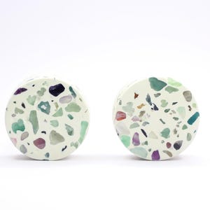 Op de afbeelding: Twee ronde onderzetters in gebroken wit met een terrazzo-ontwerp. De onderzetters hebben een gespikkeld patroon van kleurrijke chips in groen, paars, blauw en rood. Ze staan tegen een witte achtergrond.