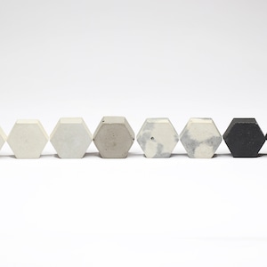 May include: A row of seven hexagonal concrete objects in varying shades of white, gray, and black. The objects are arranged in a line against a white background, showcasing their geometric shape and color variations.