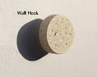 May include: A round, beige wall hook with a speckled texture. The hook is made of a stone-like material and is mounted on a light-colored wall. The text "Wall Hook" is visible in the image.
