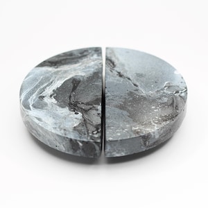 May include: Two semi-circular bookends with a marbled gray, black, and white pattern. The bookends are made of a solid material and have a smooth, polished finish. The design is abstract and modern, suitable for home decor.