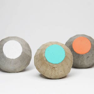 May include: Three small concrete spheres with a painted circle on each. The circles are white, turquoise, and orange.