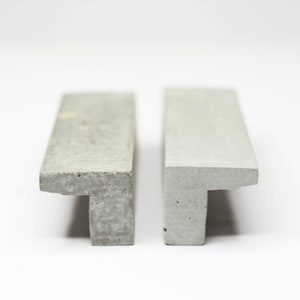 Peut inclure: Deux poignées de porte en béton avec un design en forme de T. Les poignées sont de couleur gris clair et ont une texture rugueuse.
