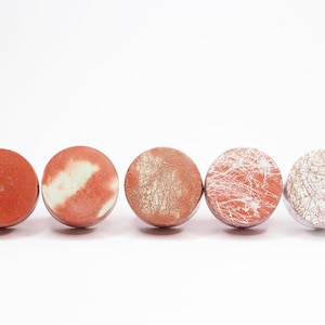 Peut inclure: Cinq boutons de meubles ronds et décoratifs dans des tons de rouge et de blanc. Les boutons présentent divers motifs de surface, notamment des couleurs unies, des motifs marbrés et des finitions texturées. Ils sont disposés en rangée sur fond blanc.