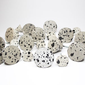 Peut inclure: Une collection de boutons en béton ronds et coniques avec un motif moucheté noir et blanc. Les boutons sont disposés sur une surface blanche.