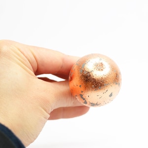 May include: A copper-colored, spherical object with a textured surface. The sphere has areas of darker discoloration, suggesting an aged or weathered appearance. The object is held in a hand against a white background.