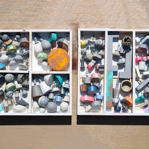 Pode incluir: Duas bandejas brancas divididas preenchidas com uma variedade de botões e puxadores coloridos. Os botões são de várias formas, tamanhos e cores, incluindo cinza, azul, rosa e amarelo. As bandejas estão em uma superfície de madeira.