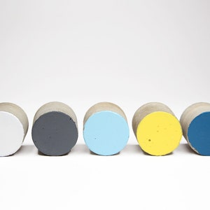 May include: Five small, round concrete cylinders painted in different colours. The cylinders are arranged in a row from left to right: white, grey, light blue, yellow, and blue.