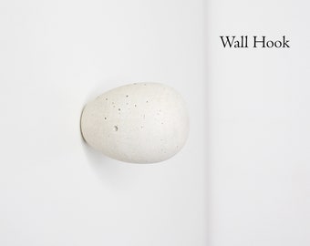 May include: A white concrete wall hook shaped like a rounded egg with a rough texture. The hook is mounted on a white wall.