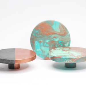 May include: Three decorative objects with a modern design. One is a gray and copper-colored disc with a short cylindrical base. Another is a round object with a marbled teal and copper pattern. The third is a small disc with a teal and copper pattern.