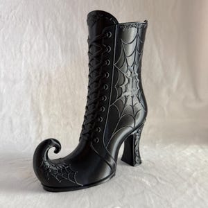 May include: A black boot-shaped decorative object featuring a curled toe, a high heel, and a lace-up front design. The exterior surface is molded with a prominent spider web pattern that extends across the shaft and the curled toe area.