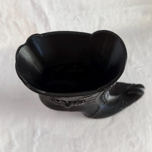 May include: A black boot-shaped container with a glossy finish, viewed from directly above. The opening is wide and oval-shaped, revealing a hollow interior. The exterior surface features molded decorative stitching and a pointed toe silhouette.