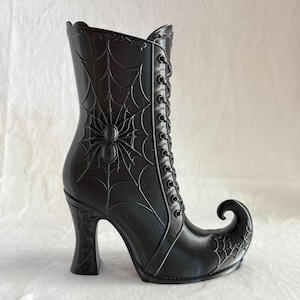 May include: A glossy black boot-shaped container featuring a raised spider and web design on the side. The boot has a high heel, a pointed curled toe, and a decorative lace-up front detail with molded eyelets extending up the shaft.
