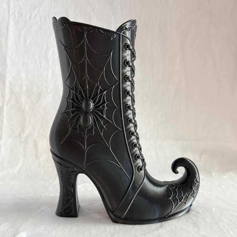 May include: A glossy black boot-shaped container featuring a raised spider and web design on the side. The boot has a high heel, a pointed curled toe, and a decorative lace-up front detail with molded eyelets extending up the shaft.