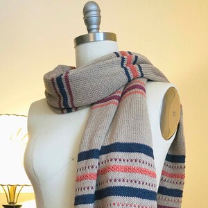 May include: A beige knitted scarf with stripes of navy blue, pink, and orange. The scarf is draped over a white dress form.