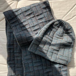 May include: A knitted scarf and beanie set in shades of blue, grey, and brown. The textured knitwear features a basketweave pattern. The scarf is rectangular, and the beanie is a close-fitting cap. The set is displayed on a cream-coloured surface.