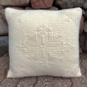 May include: A white knitted pillow with a geometric design in the center. The pillow is made of a soft, fluffy material and has a subtle sheen.