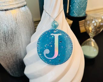 May include: A round, glittery, light blue ornament with the letter "J" in white and iridescent colors. The ornament hangs from a silver wire and a matching blue bead. The background includes a white vase and other decorative items.