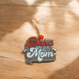 May include: A red and white wooden ornament with the text "Band Mom" and musical notes. The ornament has a silver string for hanging.