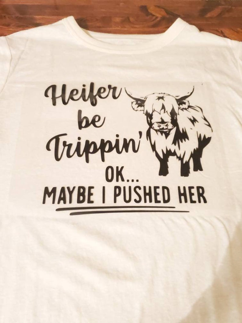 K&ouml;nnte beinhalten: Wei&szlig;es T-Shirt mit einer schwarzen Grafik einer Highland-Kuh und dem Text "Heifer be trippin' OK... MAYBE I PUSHED HER"