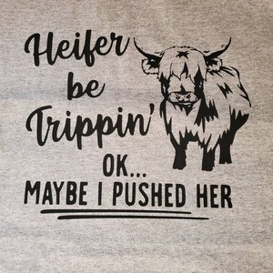 K&ouml;nnte beinhalten: Ein graues T-Shirt mit einer schwarzen Grafik einer Hochlandkuh und dem Text "Heifer be Trippin' OK... MAYBE I PUSHED HER".