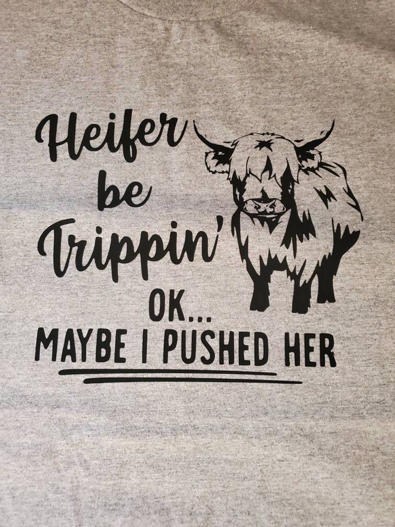 K&ouml;nnte beinhalten: Ein graues T-Shirt mit einer schwarzen Grafik einer Hochlandkuh und dem Text "Heifer be Trippin' OK... MAYBE I PUSHED HER".