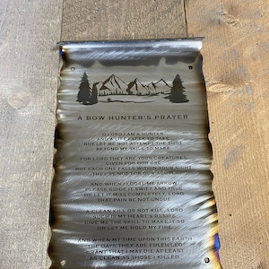 May include: A metal sign with a burnt orange and blue background featuring a poem titled "A Bow Hunter's Prayer". The poem is about the responsibility of a hunter and the respect for the animals they hunt. The sign features a silhouette of mountains and trees.