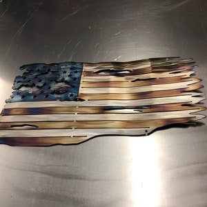 May include: A distressed American flag made of metal with a blue field of stars and red and white stripes. The flag is slightly wavy and has a distressed, burnt look.