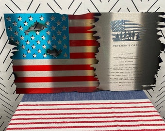 Op de afbeelding: Een Amerikaanse vlag van metaal met een verweerde look, met een blauw veld van sterren en rode en witte strepen. De vlag is gedeeltelijk verbrand en heeft aan de rechterkant een citaat over de Veteranen-Credo.