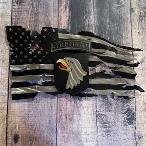 May include: A distressed metal American flag with a silver and black color scheme. The flag has a 101st Airborne Division patch with an eagle and the words "AIRBORNE" in the center.