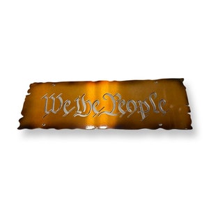 May include: A metal sign with a distressed, burnt orange finish. The sign features the words "We the People" in a silver, cursive font.