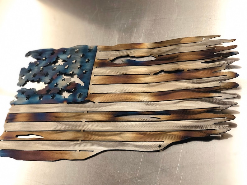 May include: A distressed American flag made of metal with a blue and white field of stars and red and white stripes. The flag is weathered and has a rustic look.