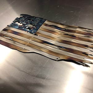 May include: A distressed metal American flag wall art with a weathered, rustic look. The flag is made of metal and has a distressed finish, giving it a vintage look. The flag is mounted on a wall and is ready to be hung.