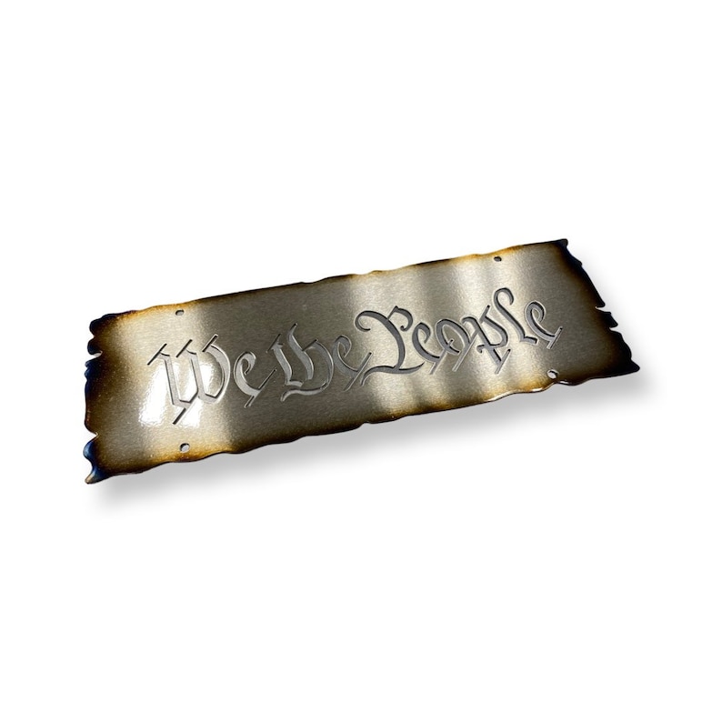 Peut inclure: Une plaque en m&eacute;tal avec un aspect vieilli et br&ucirc;l&eacute;. La plaque porte l'inscription "We the People" dans une police de caract&egrave;res stylis&eacute;e.