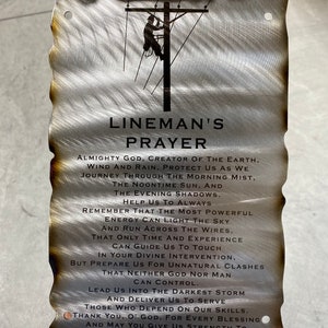 May include: A metal sign with a black silhouette of a lineman working on a power line. The sign has a weathered, distressed look and features the text "Lineman's Prayer". The prayer is a message of faith and strength for those who work in dangerous conditions.
