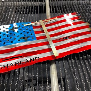 May include: A distressed American flag made of metal with the word "CHARLAND" cut out of the bottom red stripe. The flag is on a black grid surface with a ruler next to it.