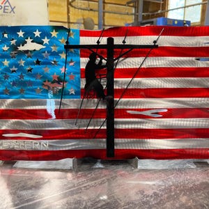 May include: A rectangular metal wall art piece featuring a distressed American flag design with red, white, and blue horizontal stripes. A black silhouette of a utility worker on a pole is centered, and the word WESTERN is embossed at the bottom.