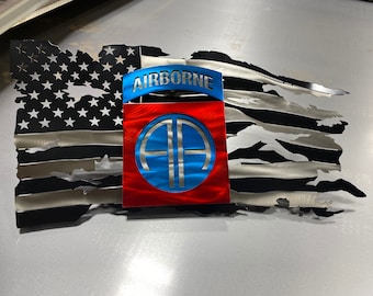 Puede incluir: Una decoración de pared de metal con una bandera estadounidense desgastada con un parche de la 101.ª División Aerotransportada de color rojo, blanco y azul. La bandera está rasgada y desgastada, lo que le da un aspecto rústico.