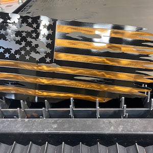 May include: Metal wall art depicting a distressed American flag. The flag features black stripes and stars with gold-colored stripes. The design has a distressed, torn appearance, with cut-out sections. The artwork is likely for indoor or outdoor display.