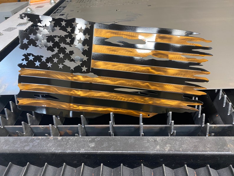 May include: Metal wall art depicting a distressed American flag. The flag features black stripes and stars with gold-colored stripes. The design has a distressed, torn appearance, with cut-out sections. The artwork is likely for indoor or outdoor display.
