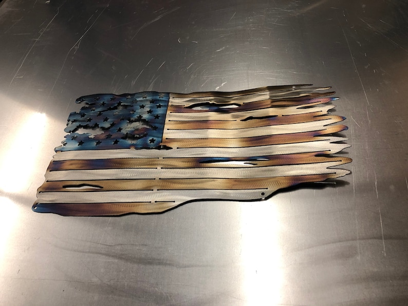 May include: A distressed American flag made of metal with a blue field of stars and red and white stripes. The flag is slightly wavy and has a distressed, burnt look.