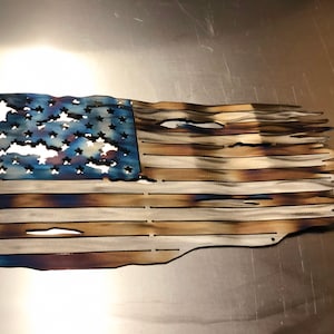 May include: A distressed American flag made of metal with a blue field of stars and red and white stripes. The flag has a weathered, burnt look, giving it a rustic appearance.